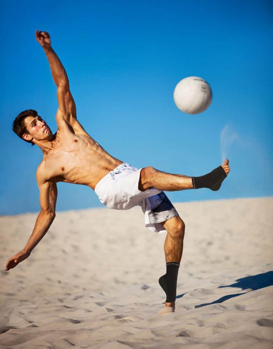 A man kicking a soccer ball in the sand with his Dr. Arthritis Copper Infused Foot Sleeve (Pair) condition unaffected.
