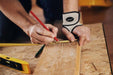 Person marking a measurement on a wooden board with a pencil, wearing a Dr. Arthritis Copper Lined Wrist Support, with a level tool nearby.