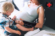 A pregnant woman experiencing wrist pain wears a Dr. Arthritis Carpal Tunnel Bundle wrist brace and sits on a couch, talking on the phone while a young boy interacts with her.
