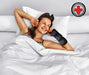 A smiling woman wearing a Carpal Tunnel Bundle wrist brace from Dr. Arthritis lying comfortably in bed with her hands behind her head on white pillows.