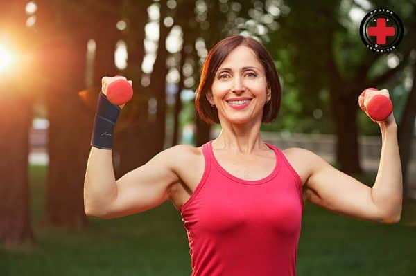 Mature woman smiling, flexing biceps with small dumbbells in a park at sunset, wearing a red tank top and Dr. Arthritis Carpal Tunnel Bundle wrist weights to prevent wrist pain, greenery in the background.