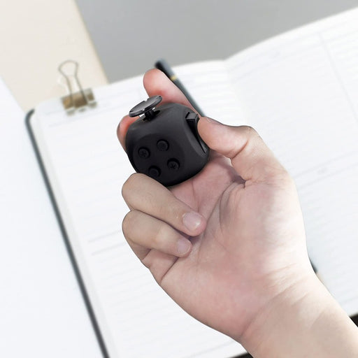 A hand holding a small black Button Fidget Cube for Adults and All Ages in front of a notebook. (Dr. Arthritis)